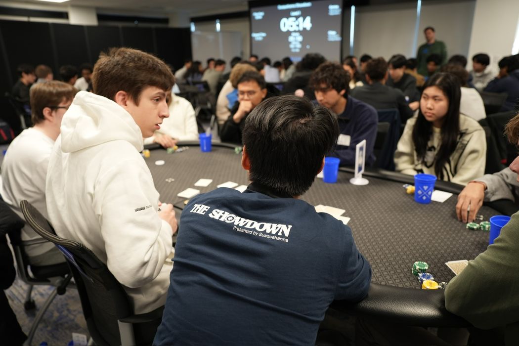 Players around a felt table with chips and cards at the Susquehanna Showdown, a tournament clock on the screen behind them