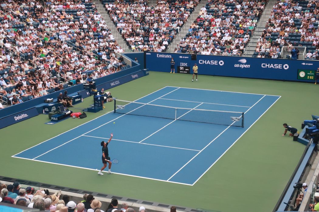Coleman Wong tosses the ball to serve on the blue court of Louis Armstrong Stadium, with a packed crowd around the court