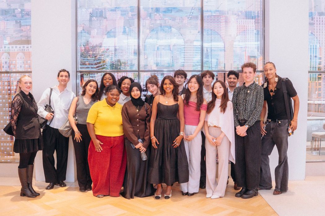 A group of program participants pose together in front of a tall window with a painted city scene