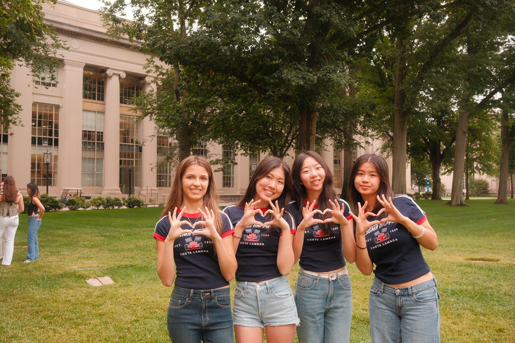 Four Sigma Kappa members in matching Theta Lambda shirts make heart hands on a lawn in front of a columned MIT building